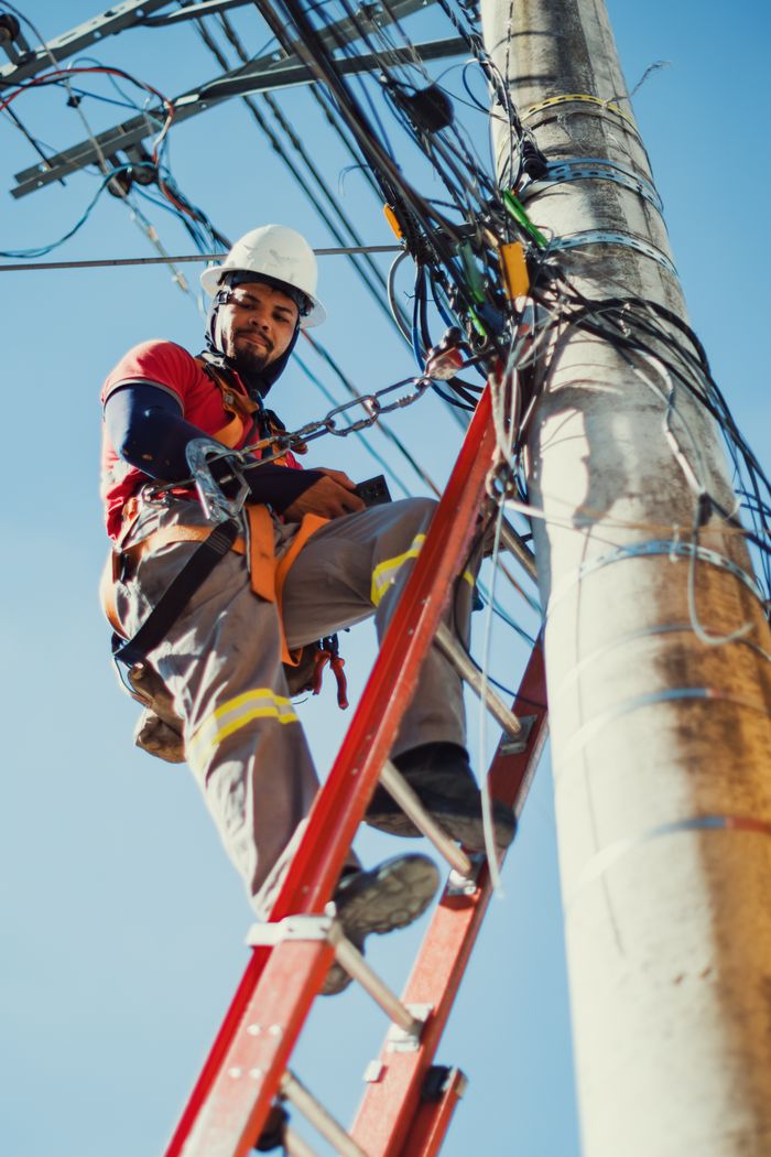 Services Technician climbing a ladder to repair power lines on a utility pole under clear blue sky.